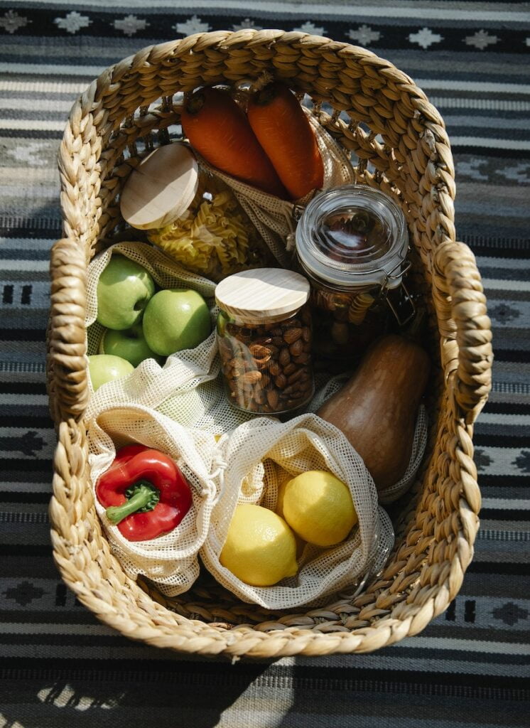From above of wicker basket with vegetables and fruits placed in eco bags and glass jars with products in sunny day